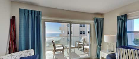A modern living room with a sofa, a chair, a small table, and a lamp. There is a sliding glass door leading to a balcony with a view of buildings and the sea.