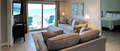 A living room with a sofa, a coffee table, and a TV. There is a sliding glass door leading to a balcony with chairs.