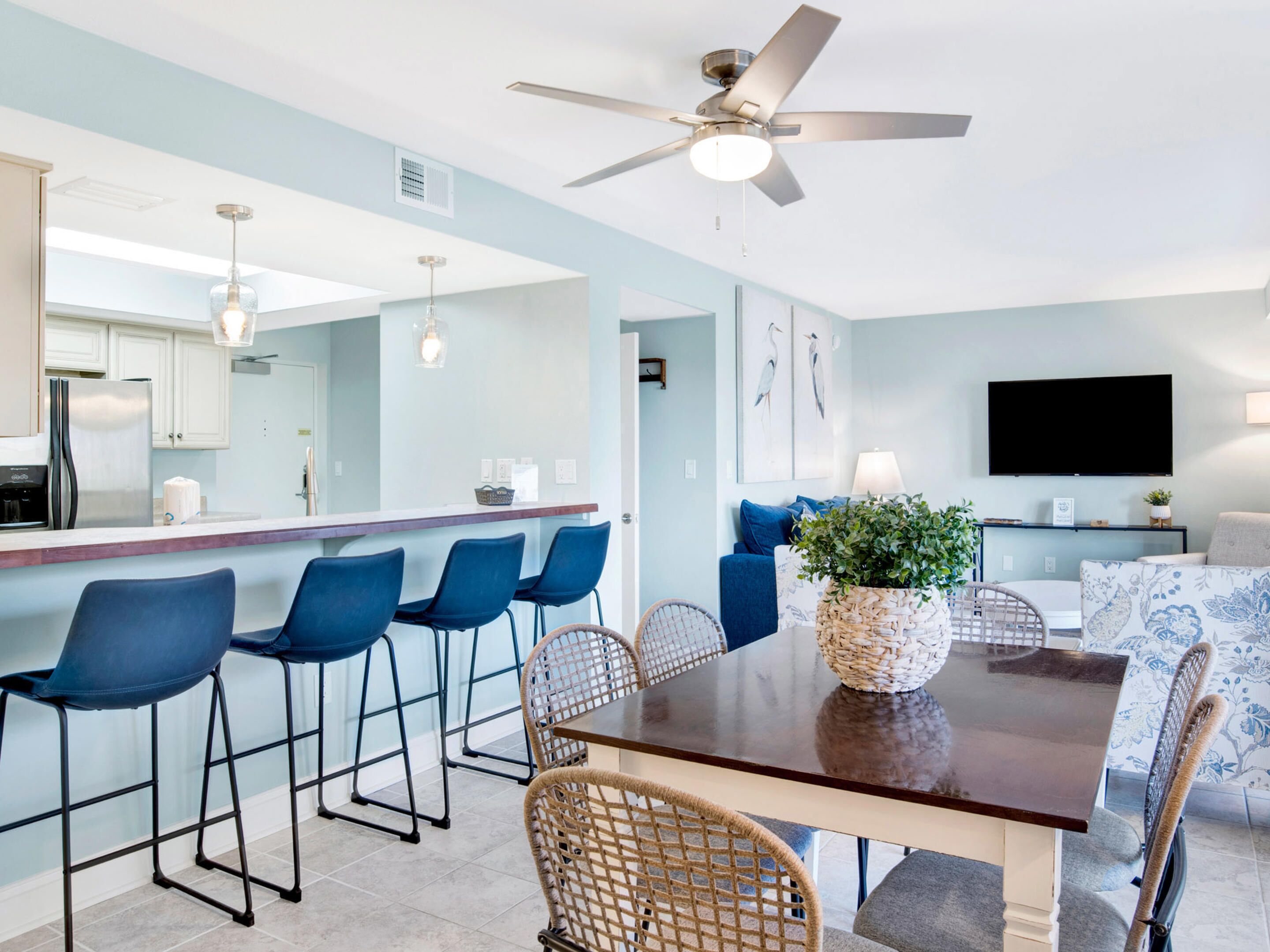 A modern kitchen with white cabinets, a bar area with blue chairs, a dining table with a woven centerpiece, and a ceiling fan.