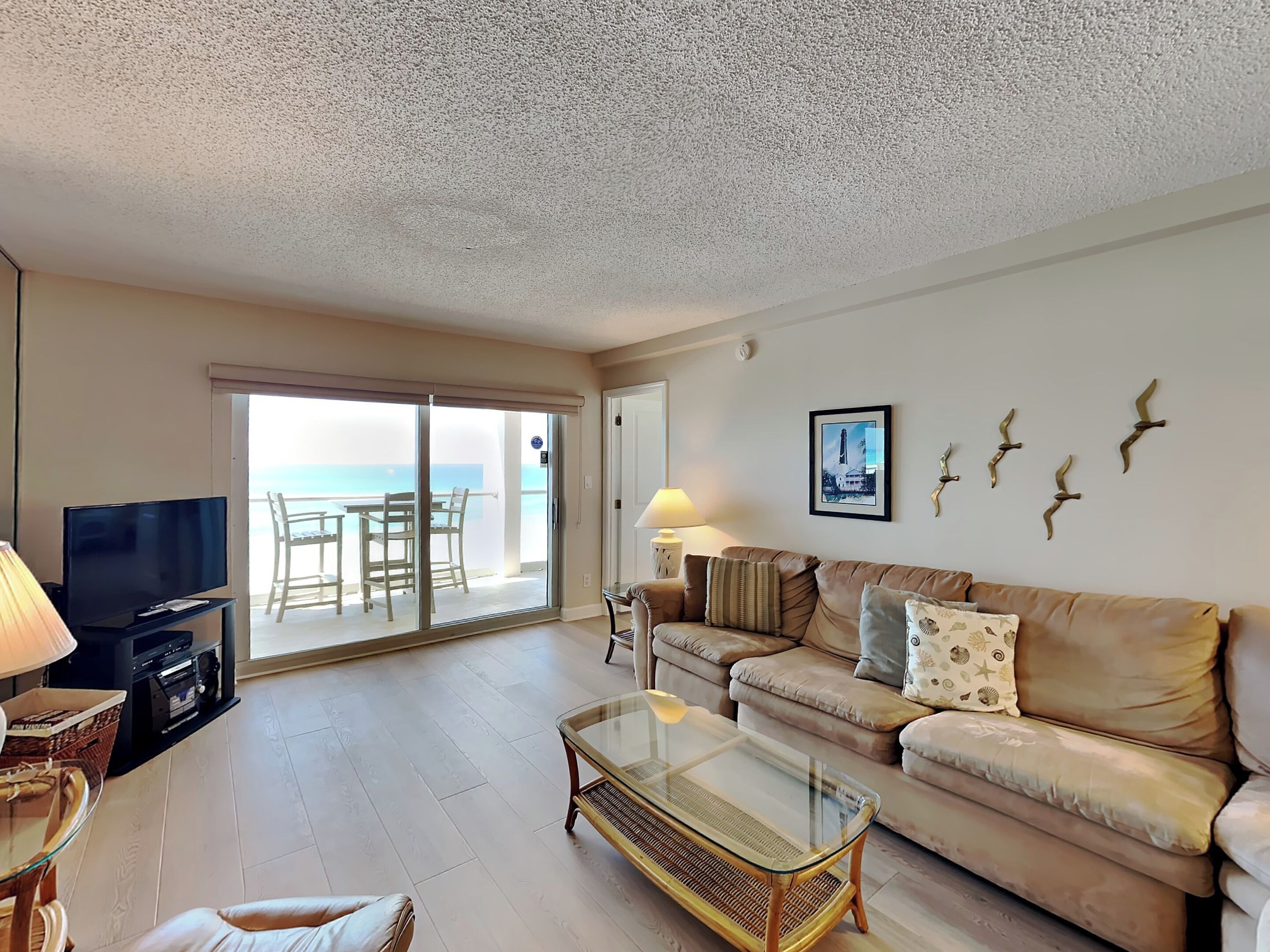 Una sala de estar con televisor, sofá, mesa de centro de vidrio y un balcón con vista al mar.