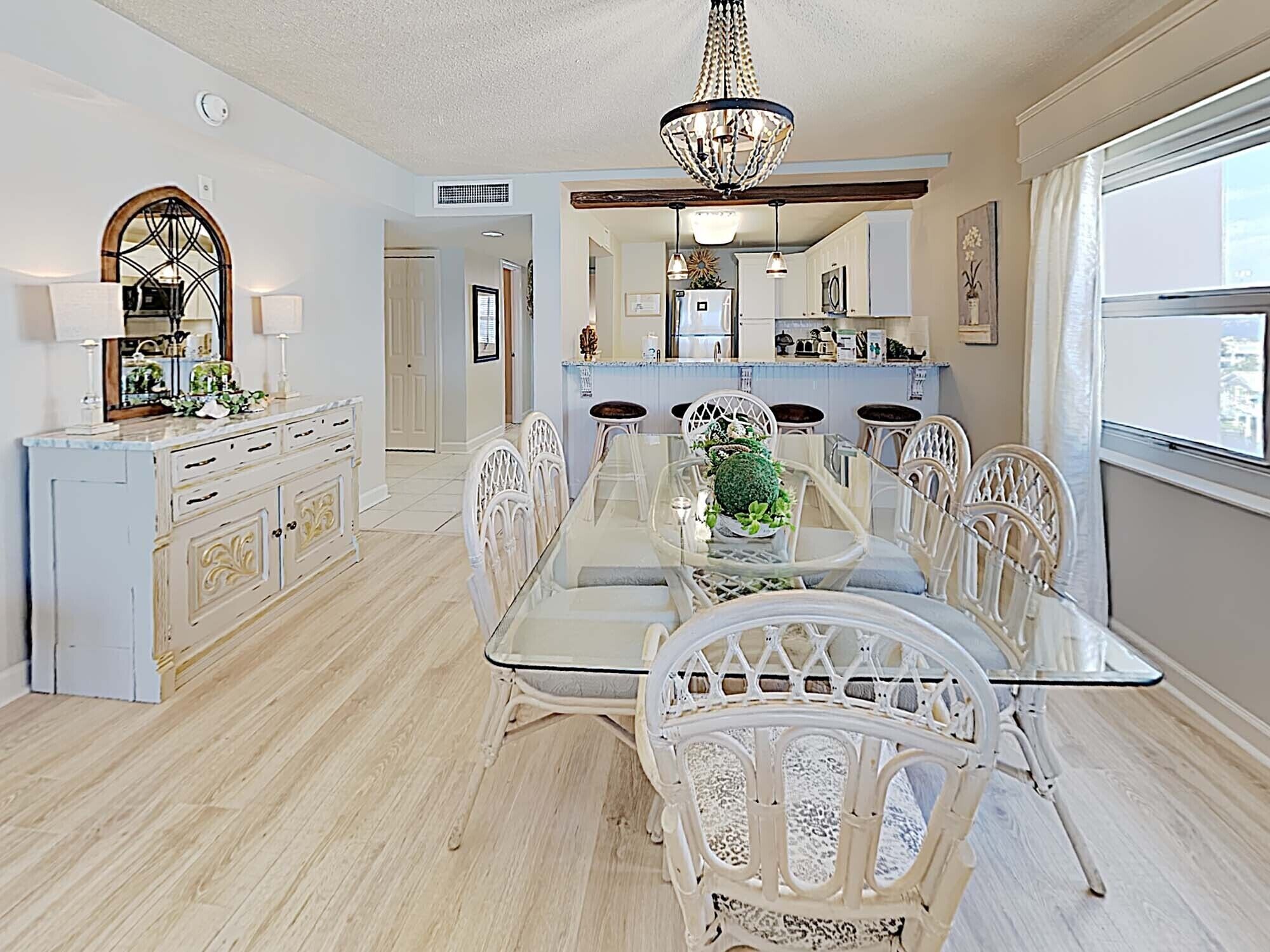 A dining area with a glass table and white chairs, a chandelier, and a view of the kitchen in the background.