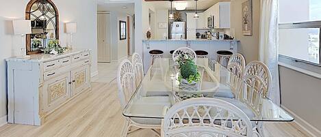 A dining area with a glass table and white chairs, a chandelier, and a view of the kitchen in the background.