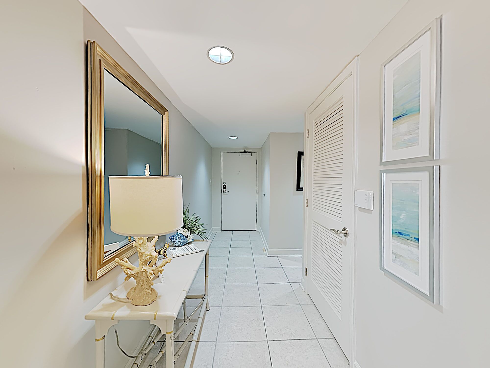 Um corredor com piso de azulejos brancos, um lavatório branco com espelho em moldura dourada, uma porta branca com janela e obras de arte emolduradas na parede.