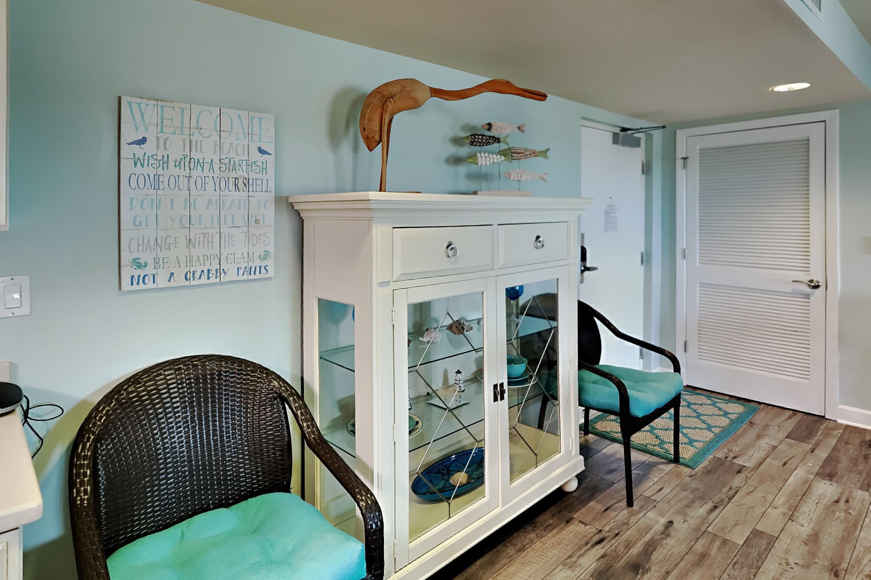 Una stanza con un armadietto bianco con ante in vetro, due poltrone in vimini con cuscini color turchese, una decorazione a forma di pesce di legno e un cartello di benvenuto appeso al muro.