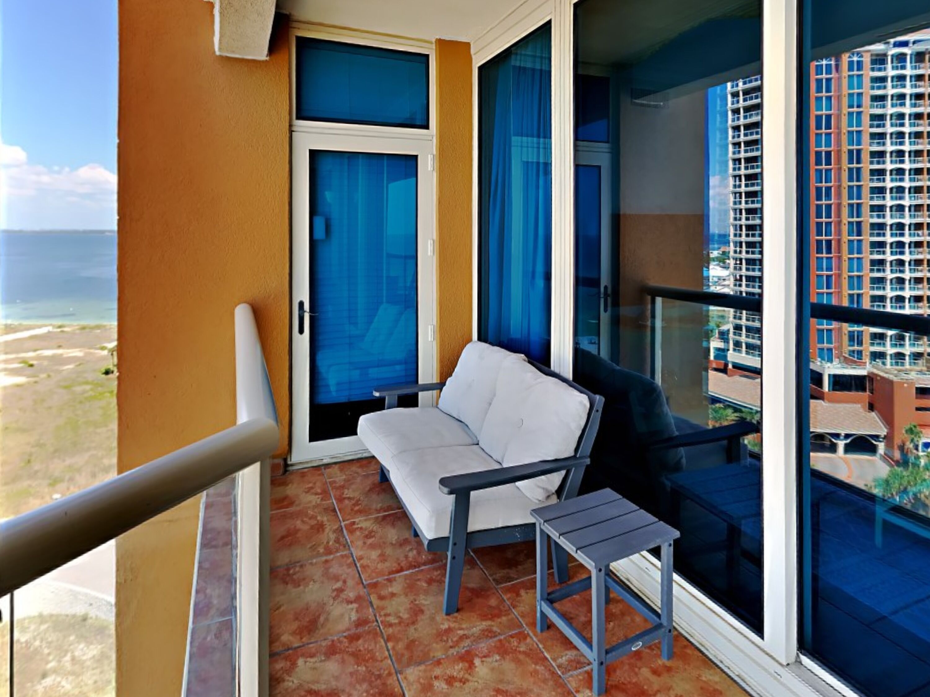 A balcony with a white sofa, a small table, and a view of a building and beach.