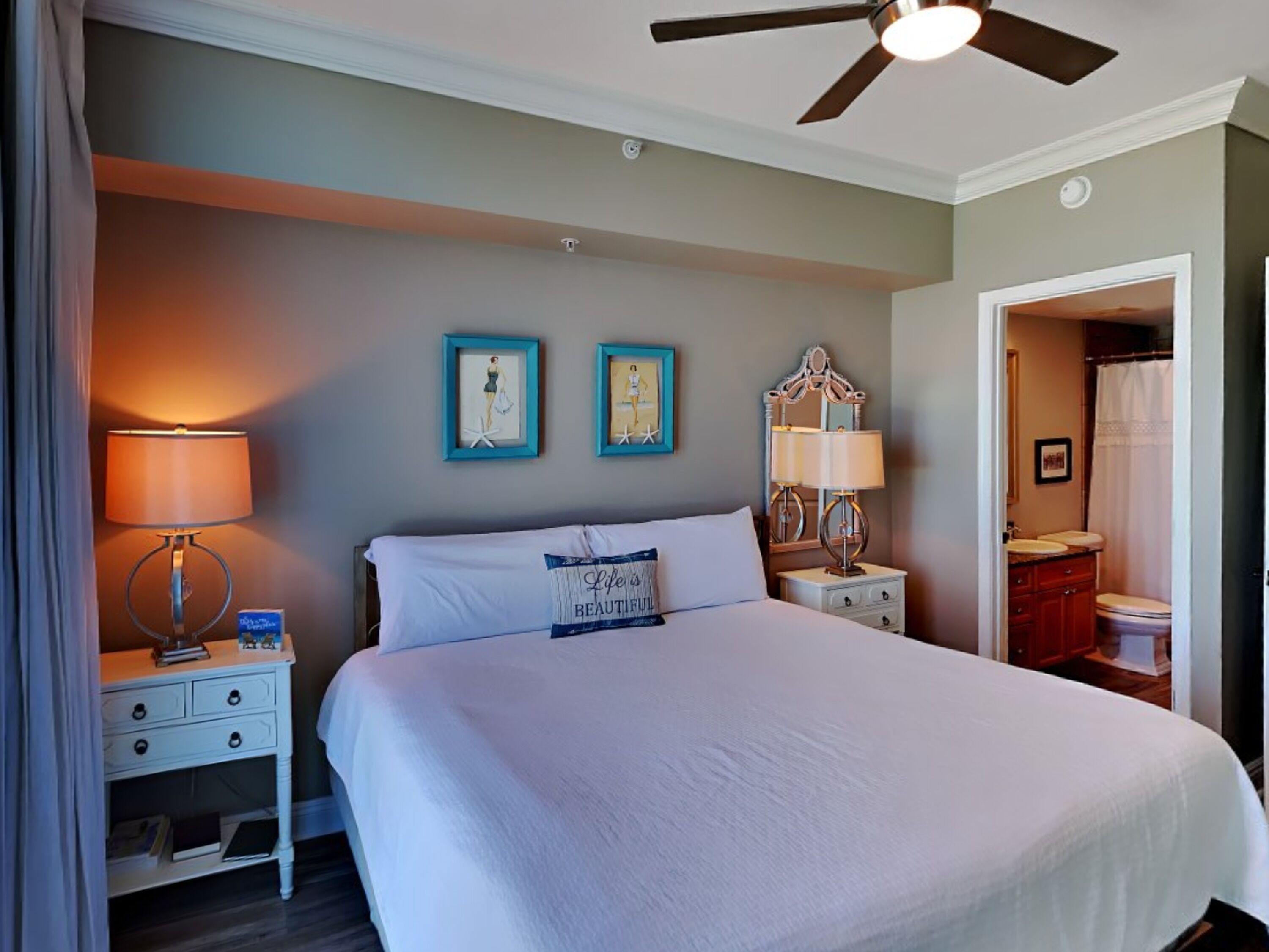 A hotel room with a large bed, two bedside tables with lamps, a ceiling fan, and a bathroom visible through an open door.