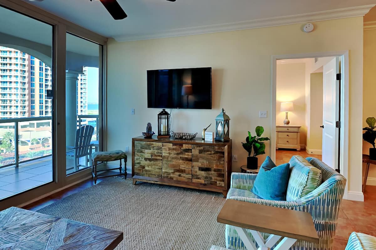 A spacious living room with a large window, a ceiling fan, a flat-screen TV, a wooden cabinet, and a patterned sofa.