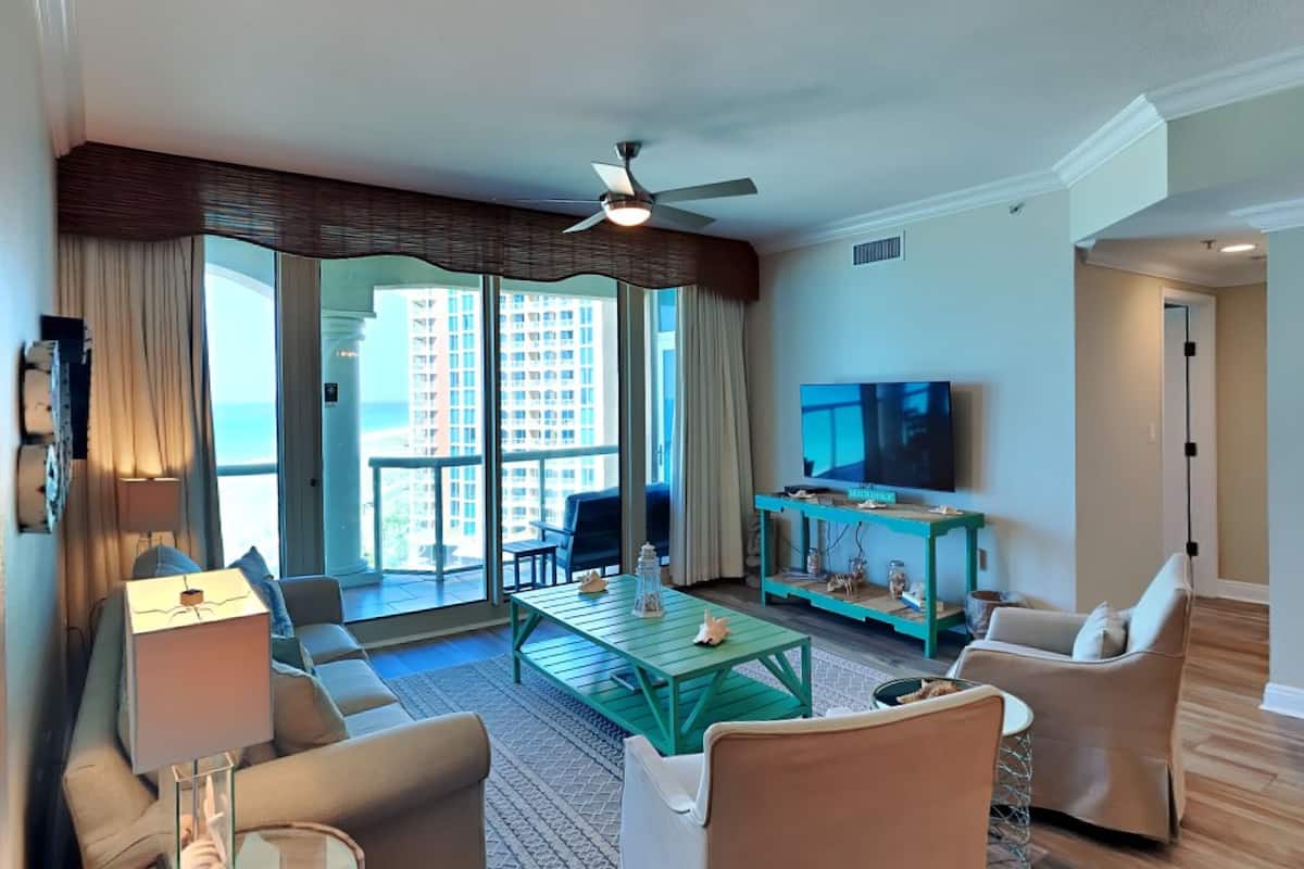 A spacious living room with a large TV, comfortable seating, a balcony with a view of tall buildings, and a ceiling fan.