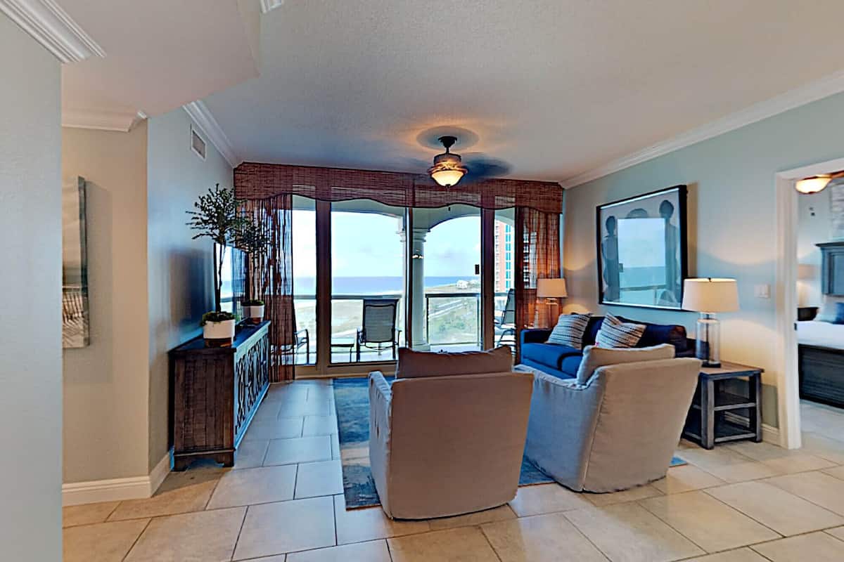 A spacious living room with a large window offering a view of the ocean, a ceiling fan, and a dining area with a view.