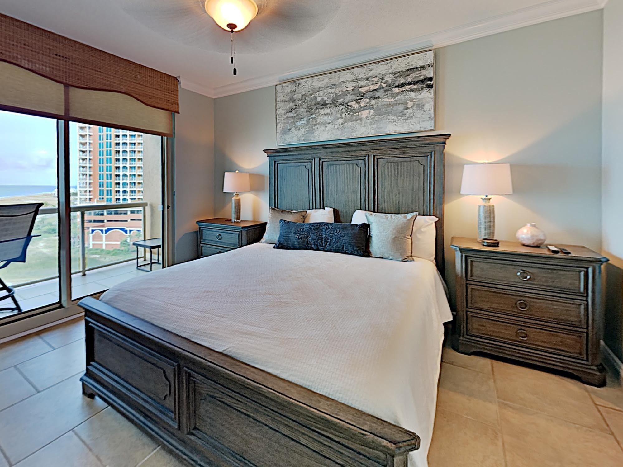 A bedroom with a large bed, two bedside tables, a nightstand, a view of the ocean, and a balcony.