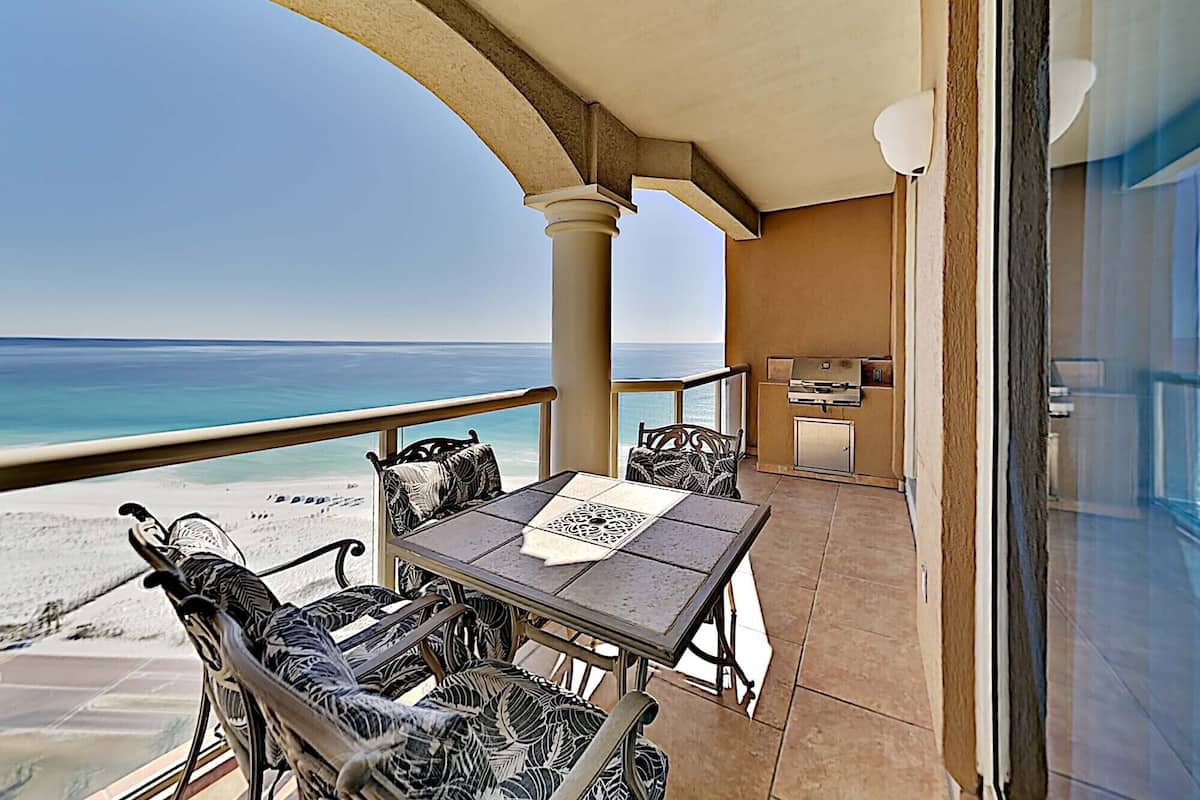A balcony with a view of the beach and ocean, a table with chairs, and a grill area.