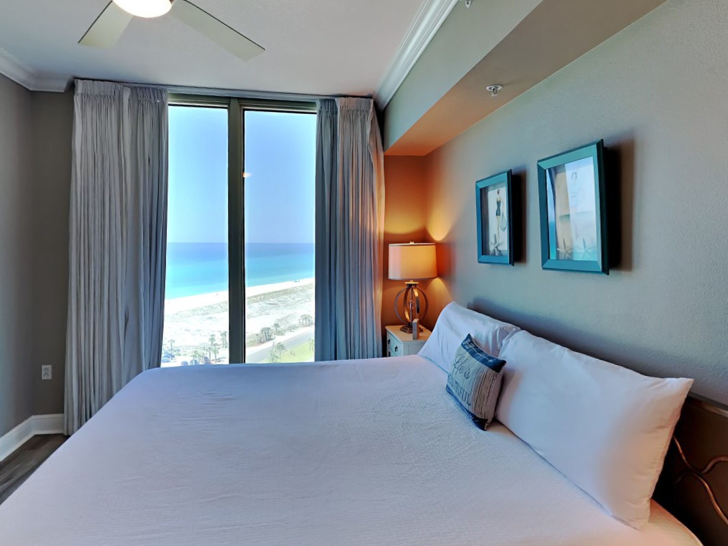 A hotel room with a large bed, a nightstand, a lamp, and a view of the beach.