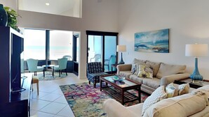 A spacious living room with a large window, a ceiling fan, a sofa, a coffee table, and a TV. - Palm Beach Club by Southern Vacation Rentals (Pensacola Beach)