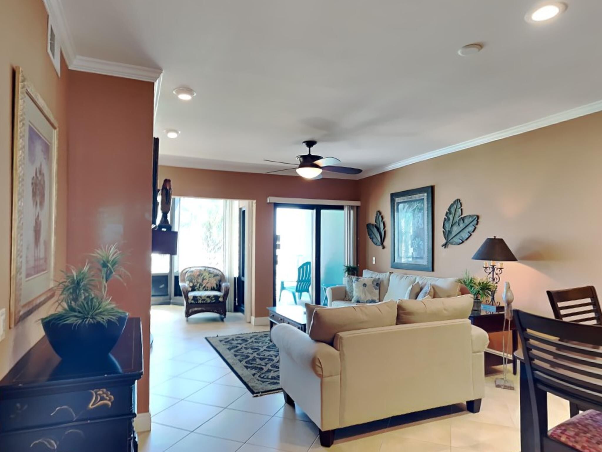 A living room with a beige sofa, a coffee table, and a ceiling fan. There is a large window and a door leading to another room.