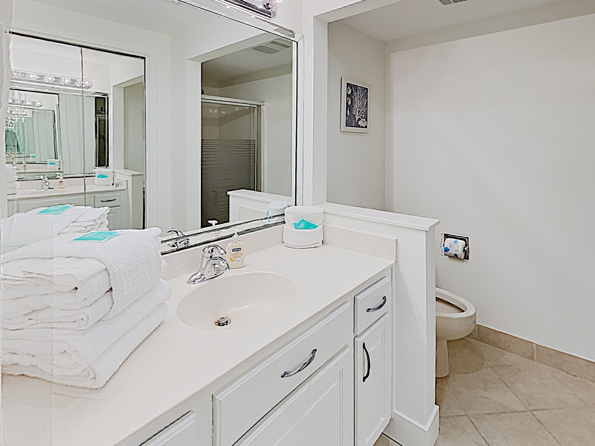 A modern bathroom with a white sink, vanity, and a large mirror. There is a towel rack with folded towels and a toilet visible in the corner.