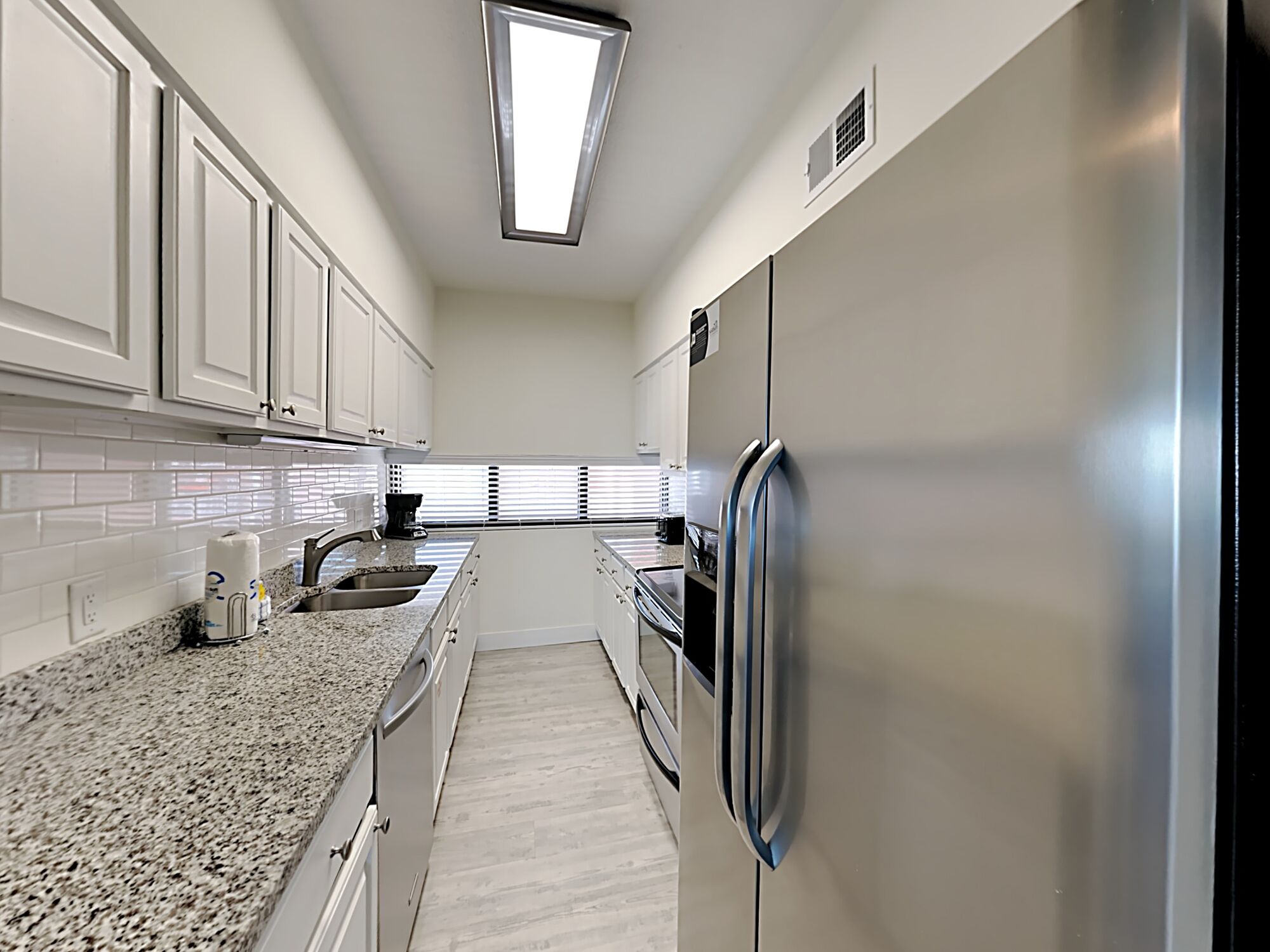 A modern kitchen with white cabinets, a stainless steel refrigerator, granite countertops, and a window with blinds.