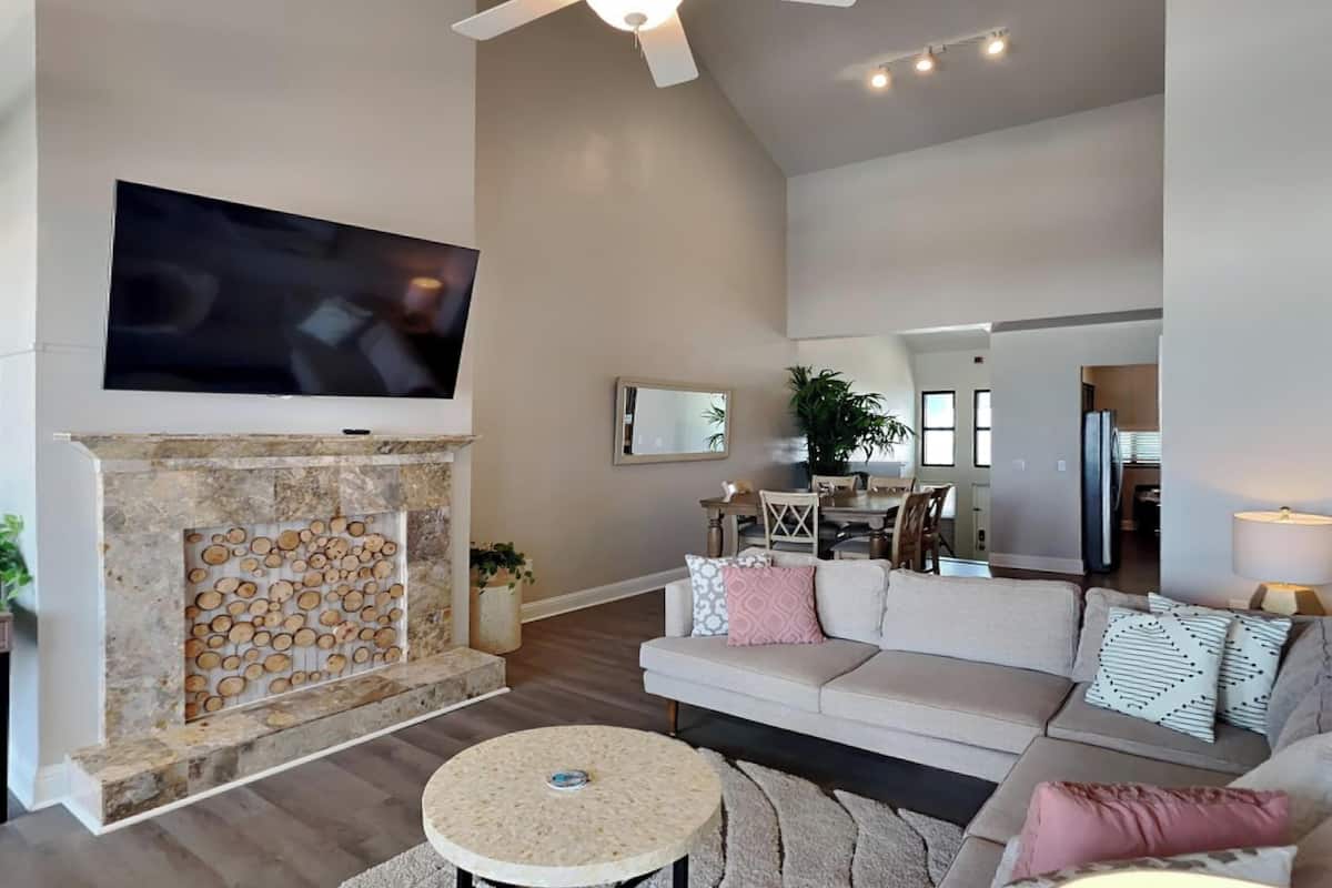 A modern living room with a stone fireplace, a flat-screen TV, a sofa with patterned cushions, and a dining area in the background.