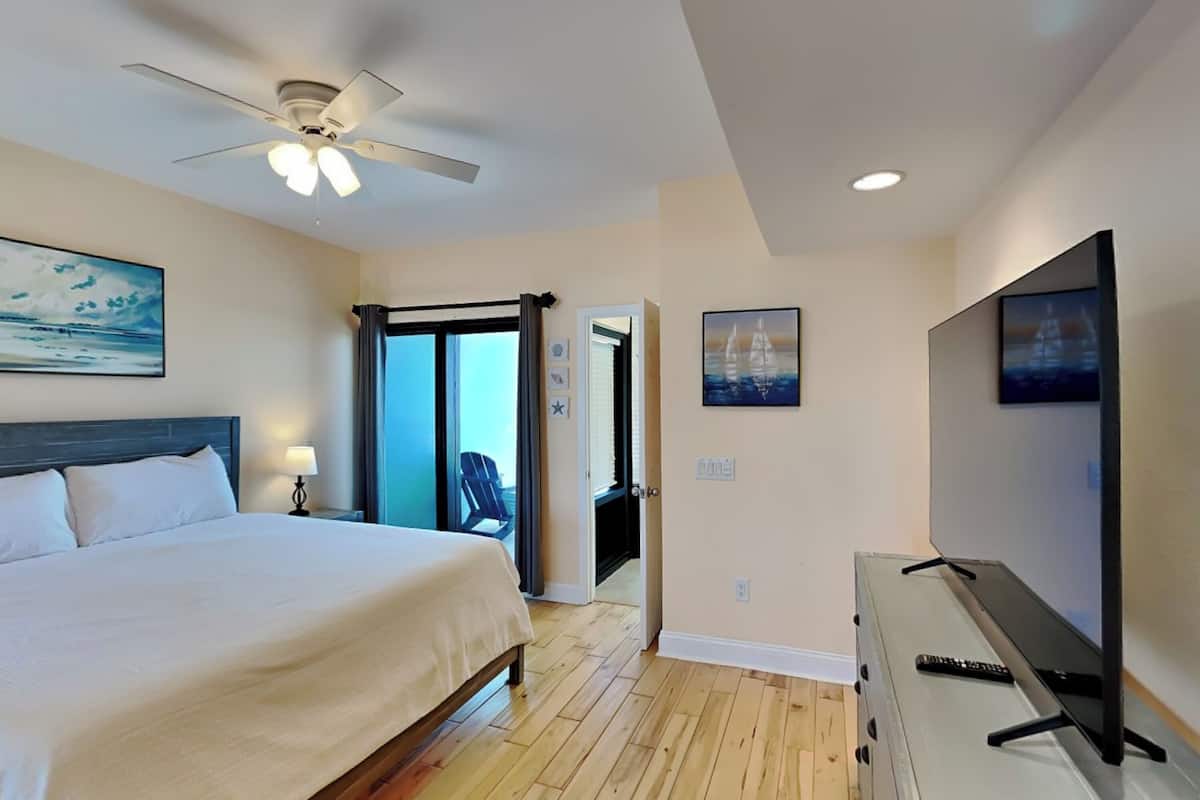 A hotel room with a bed, ceiling fan, artwork, and a television.