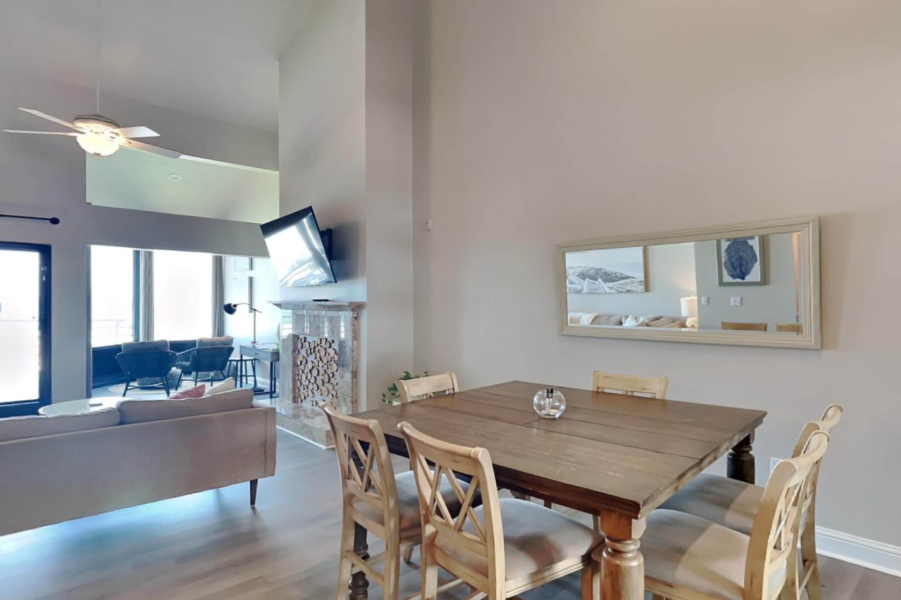 A spacious living room with a large wooden dining table, comfortable seating, a ceiling fan, and a large mirror on the wall.