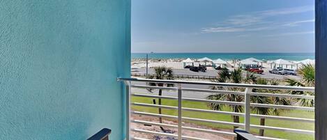 A balcony with two lounge chairs, a view of the beach, and beach umbrellas in the distance.