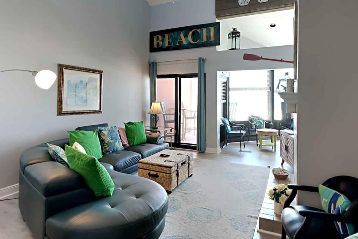 A modern living room with a sofa, a coffee table, and a bookshelf. There is a large window with a view of the outdoors.