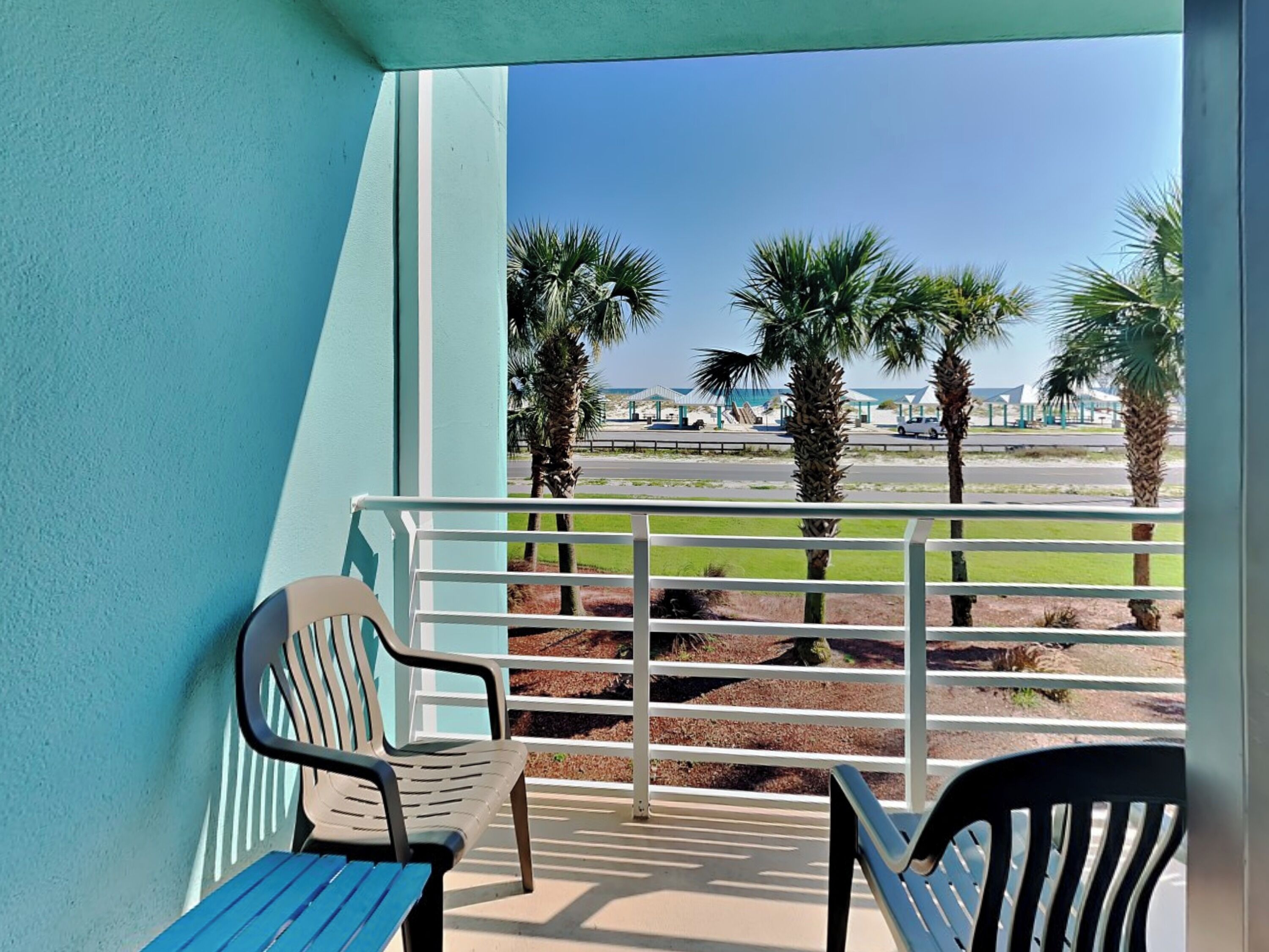 Ein Balkon mit zwei Stühlen und einem Tisch, der auf einen Strand mit Palmen und einen Parkplatz blickt.