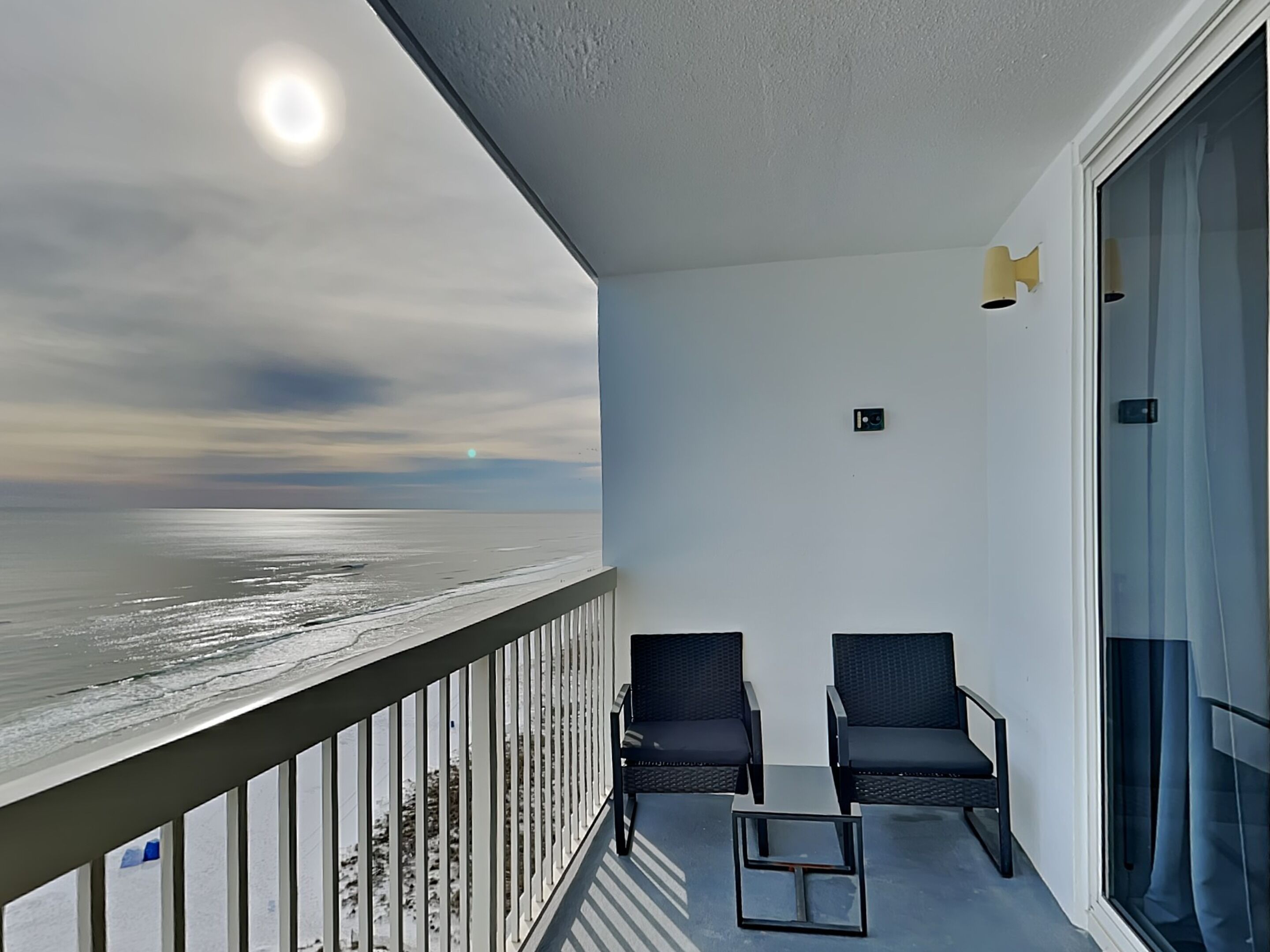 A balcony with two chairs and a small table, overlooking a beach with the ocean and a cloudy sky.