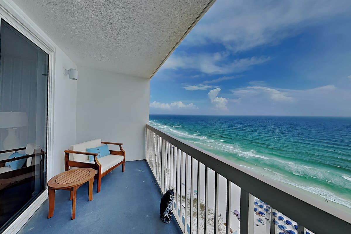 A balcony with a view of the ocean, a white sofa, a wooden table, and a black dog.
