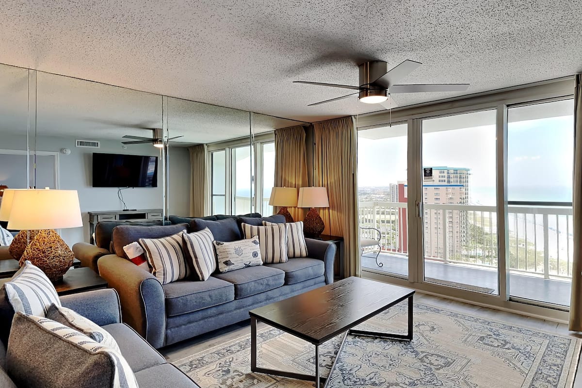 A modern hotel room with a large window, a sofa, a coffee table, and a ceiling fan.