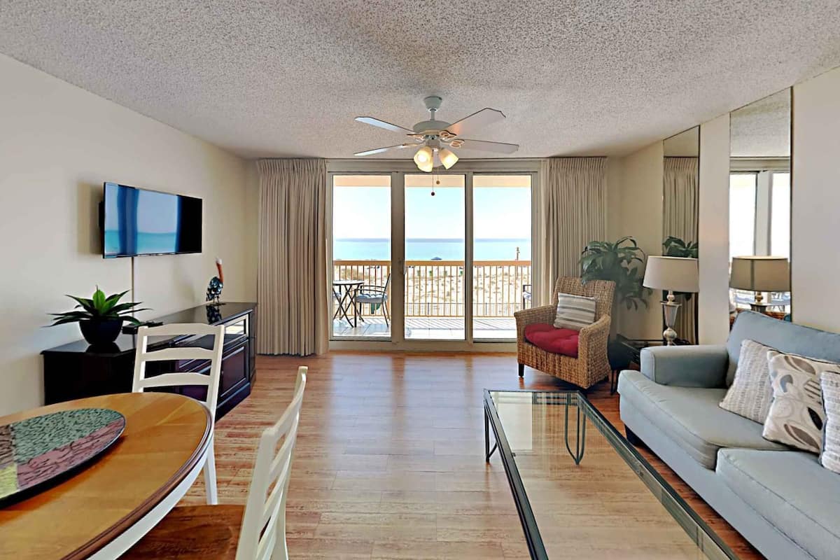 A living room with a television, a dining table, a sofa, and a balcony with a view of the beach.