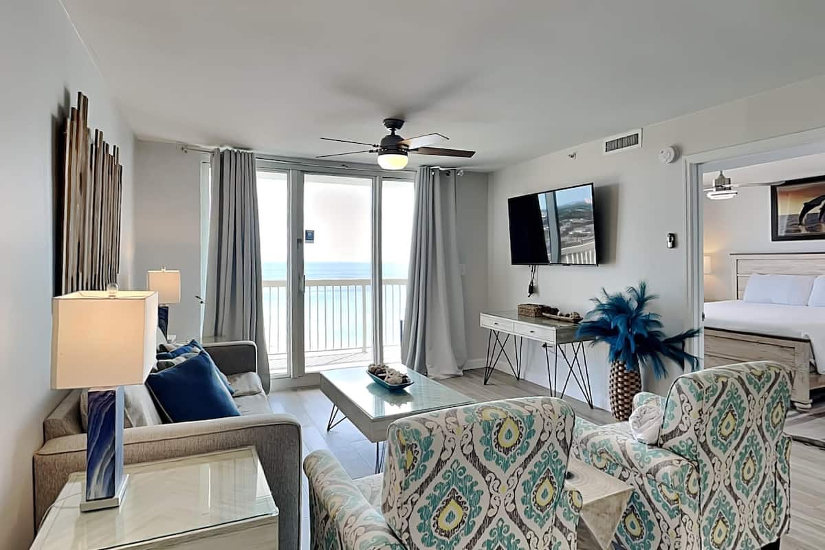 A hotel room with a balcony, a bed, a TV, a desk, and patterned armchairs.