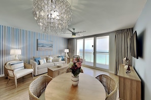 A modern living room with a dining table, wicker chairs, a sofa, and a large window with a view of the sea.