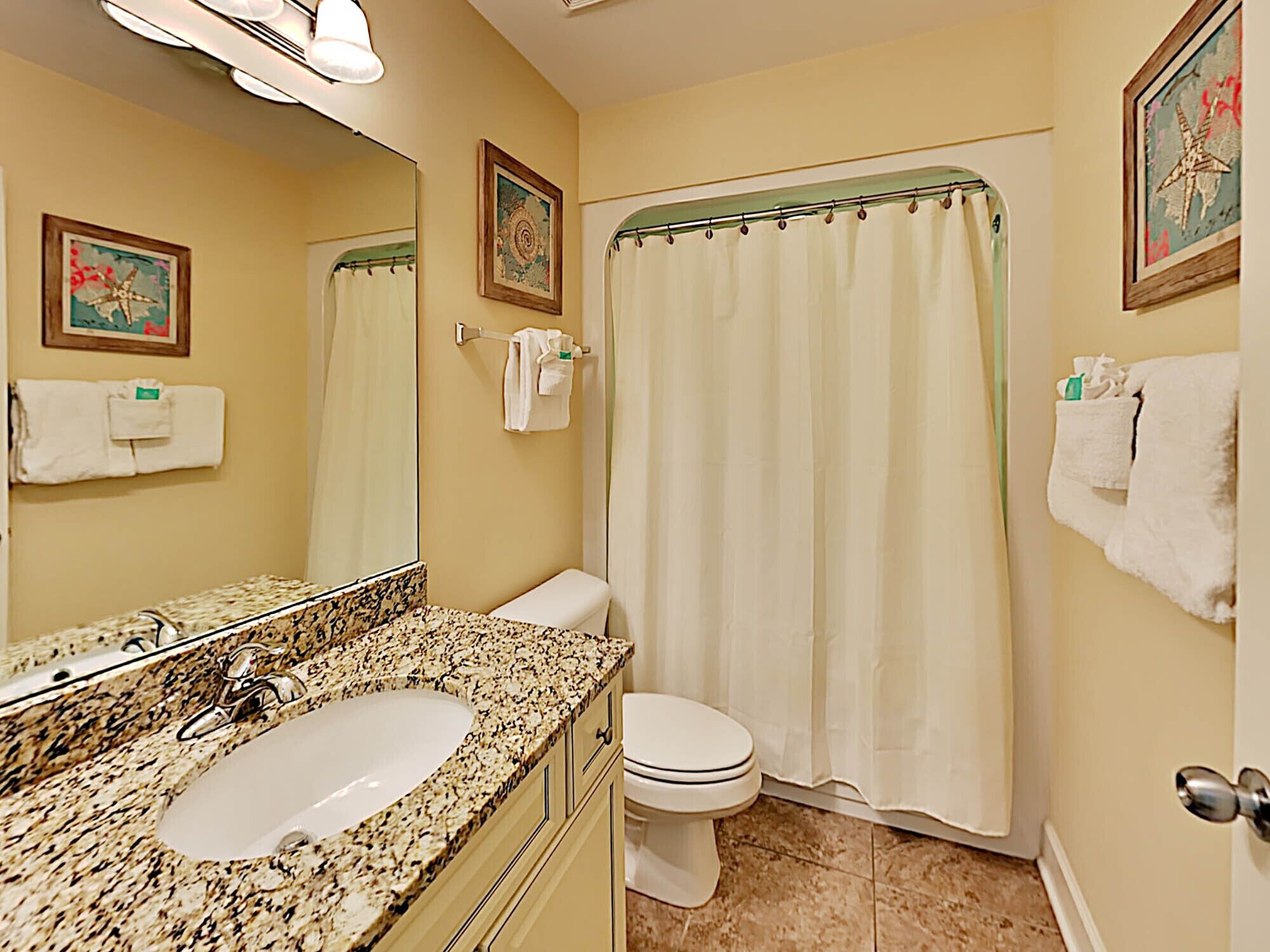 A bathroom with a granite countertop sink, a toilet, a shower with a curtain, and two framed pictures on the walls.