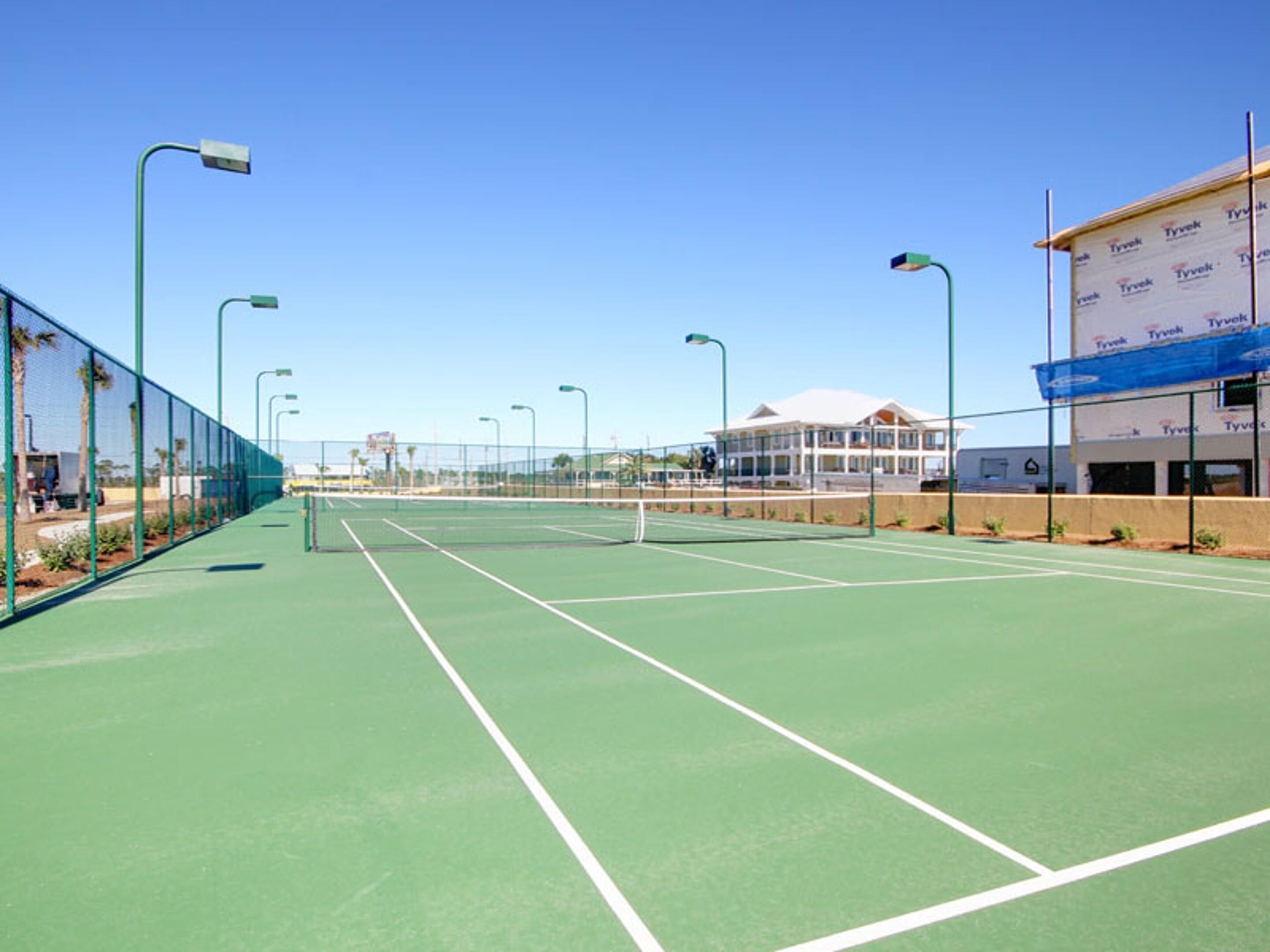 A tennis court with green flooring, surrounded by a fence and street lamps.