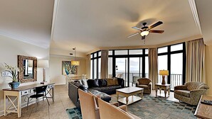 A spacious living room with a large sofa, a coffee table, and a dining area with chairs. There is a ceiling fan, large windows, and a view of the outdoors. - The Oasis at Orange Beach by Southern Vacation Rentals (Orange Beach)