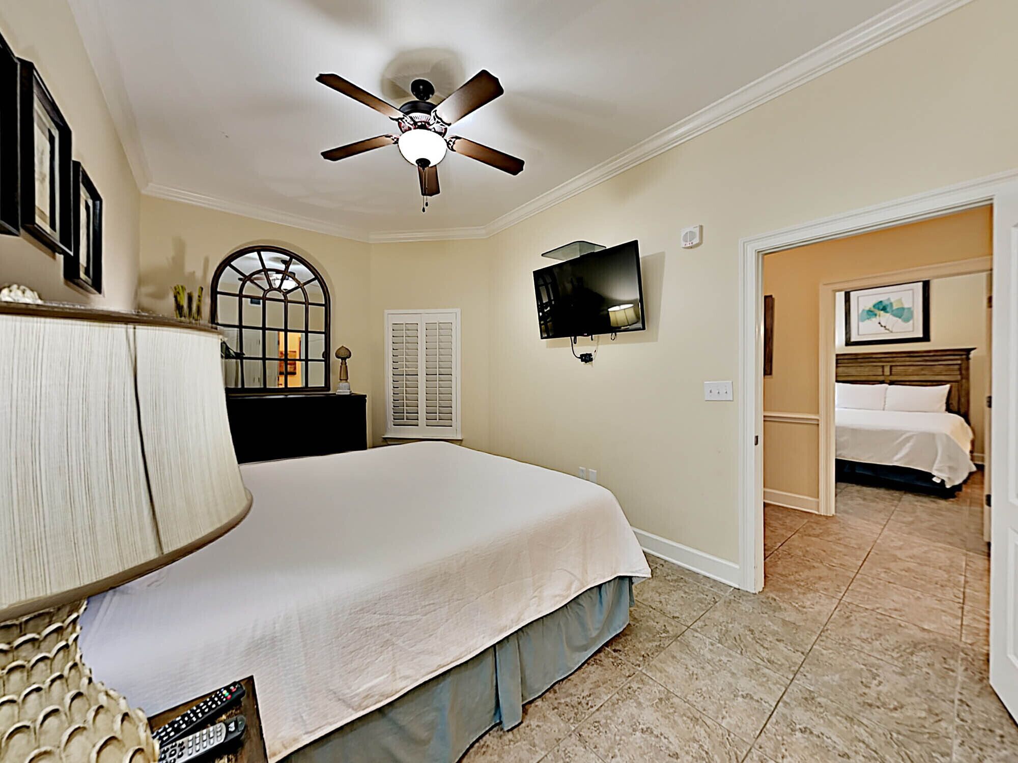 A bedroom with a bed, a ceiling fan, a TV, a mirror, and a door leading to another room.