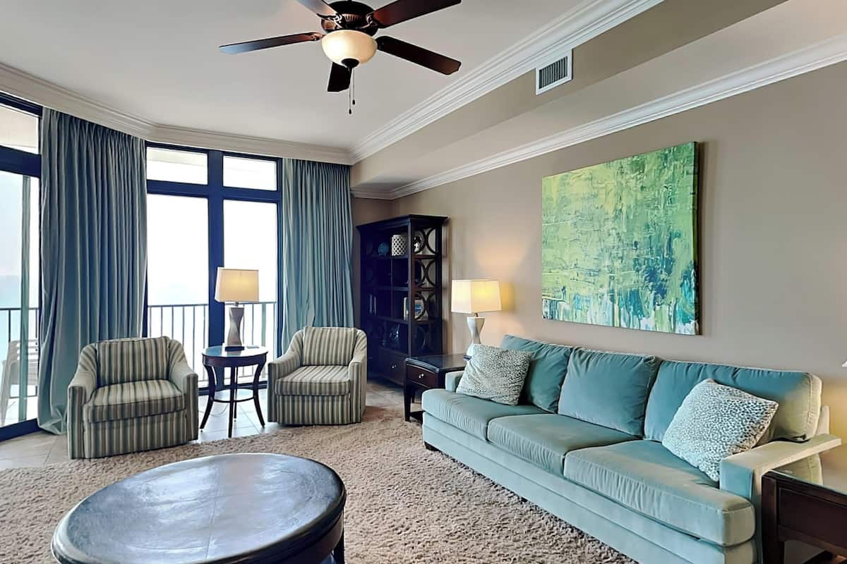Un salon comprenant un canapé, deux fauteuils, une table basse, un ventilateur de plafond et un grand tableau abstrait accroché au mur.