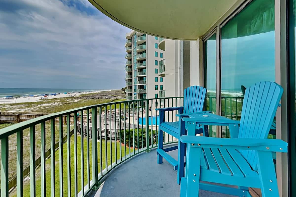 A balcony with two blue chairs, a glass railing, and a view of the beach and ocean.