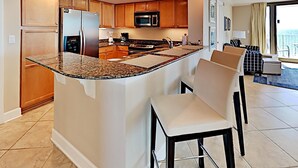 A modern kitchen with granite countertops, wooden cabinets, a central island with bar stools, and a view to the living area. - Perdido Towers by Southern Vacation Rentals (Pensacola)
