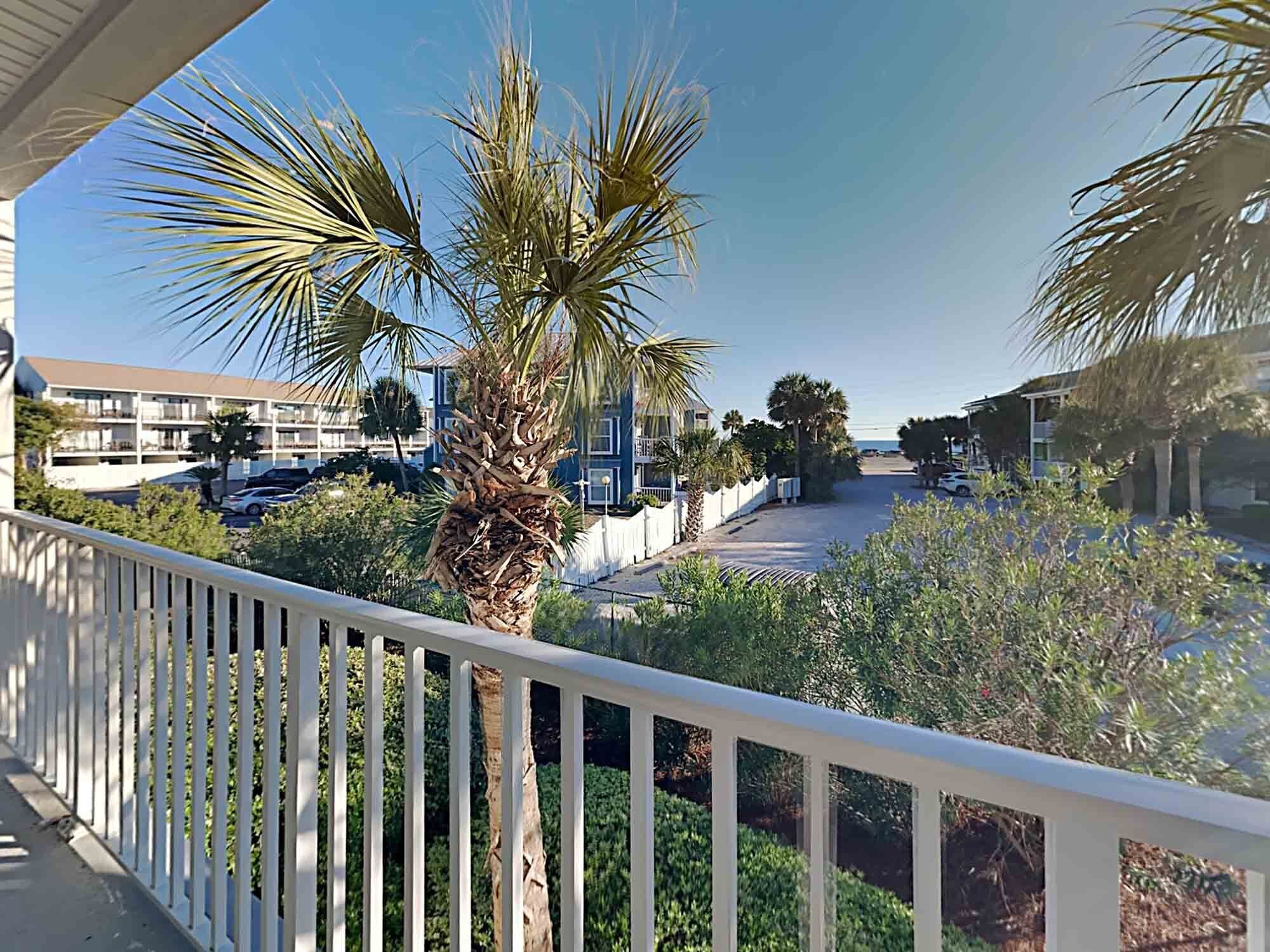 A balcony with a white railing overlooking a parking area, palm trees, and a building with multiple floors.
