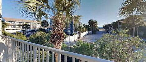 A balcony with a white railing overlooking a parking area, palm trees, and a building with multiple floors.