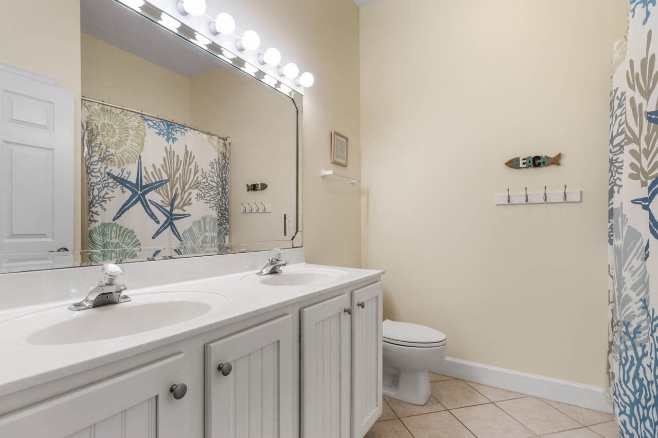 A bathroom with a double sink vanity, a large mirror with lights, a toilet, and a shower curtain with a leaf pattern.