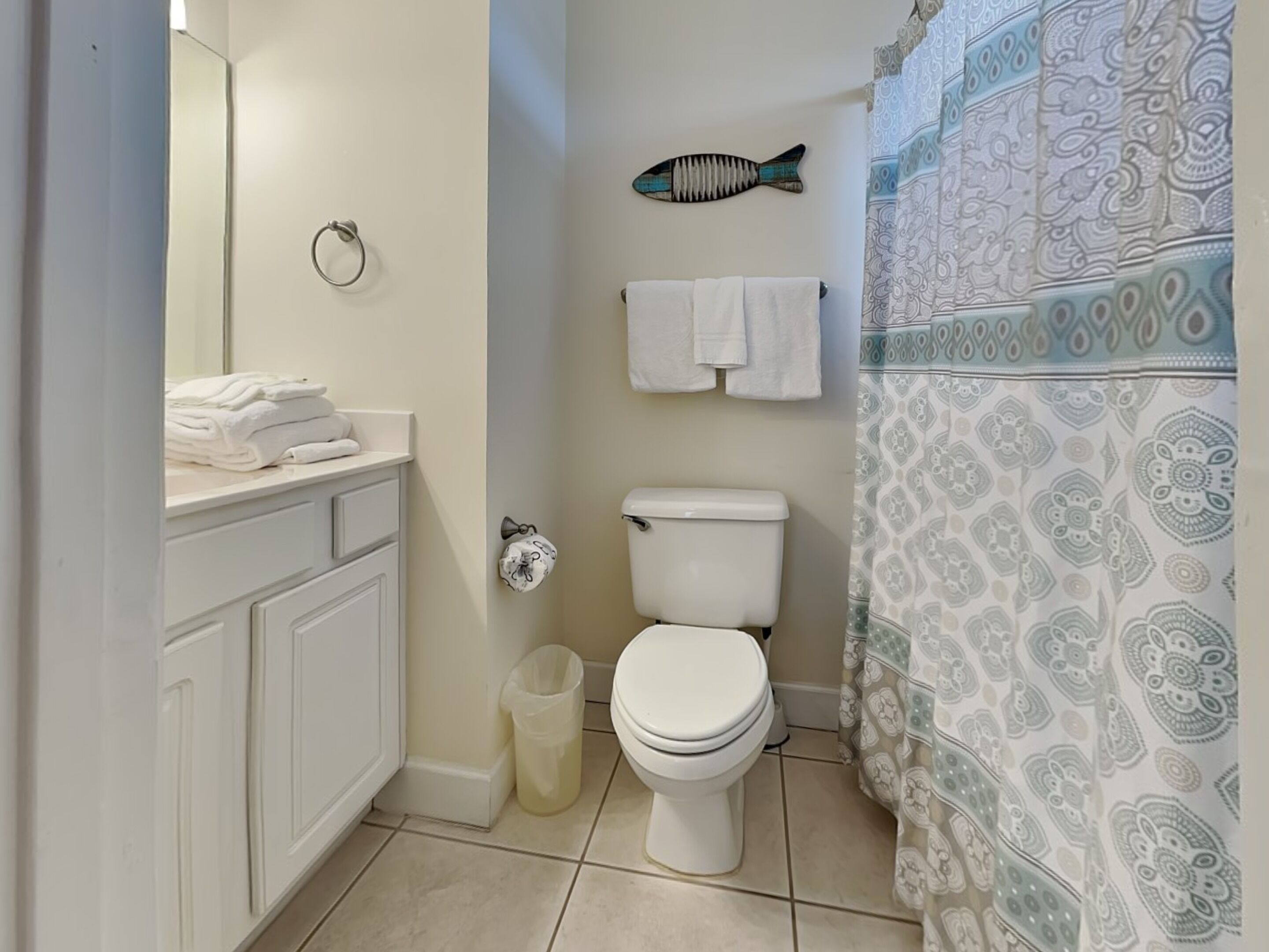 A bathroom with a toilet, a sink with a towel rack, a trash bin, and patterned shower curtains.