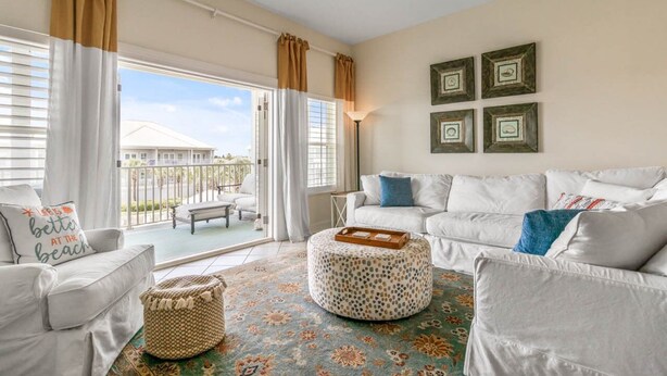 A spacious living room with a white sofa set, a round ottoman, and a patterned rug. There are two windows with curtains, a ceiling fan, and a balcony visible through the windows. - Pavilion Palms by Southern Vacation Rentals (Miramar Beach)