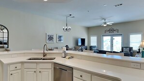 A modern kitchen with white cabinetry, a dishwasher, a sink, and a ceiling fan. There are two framed pictures on the wall and a window with blinds. - Pavilion Palms by Southern Vacation Rentals (Miramar Beach)