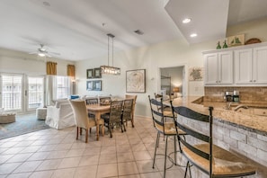 A spacious dining area with a large table and chairs, a kitchen with white cabinets and a stone countertop, and a ceiling fan.