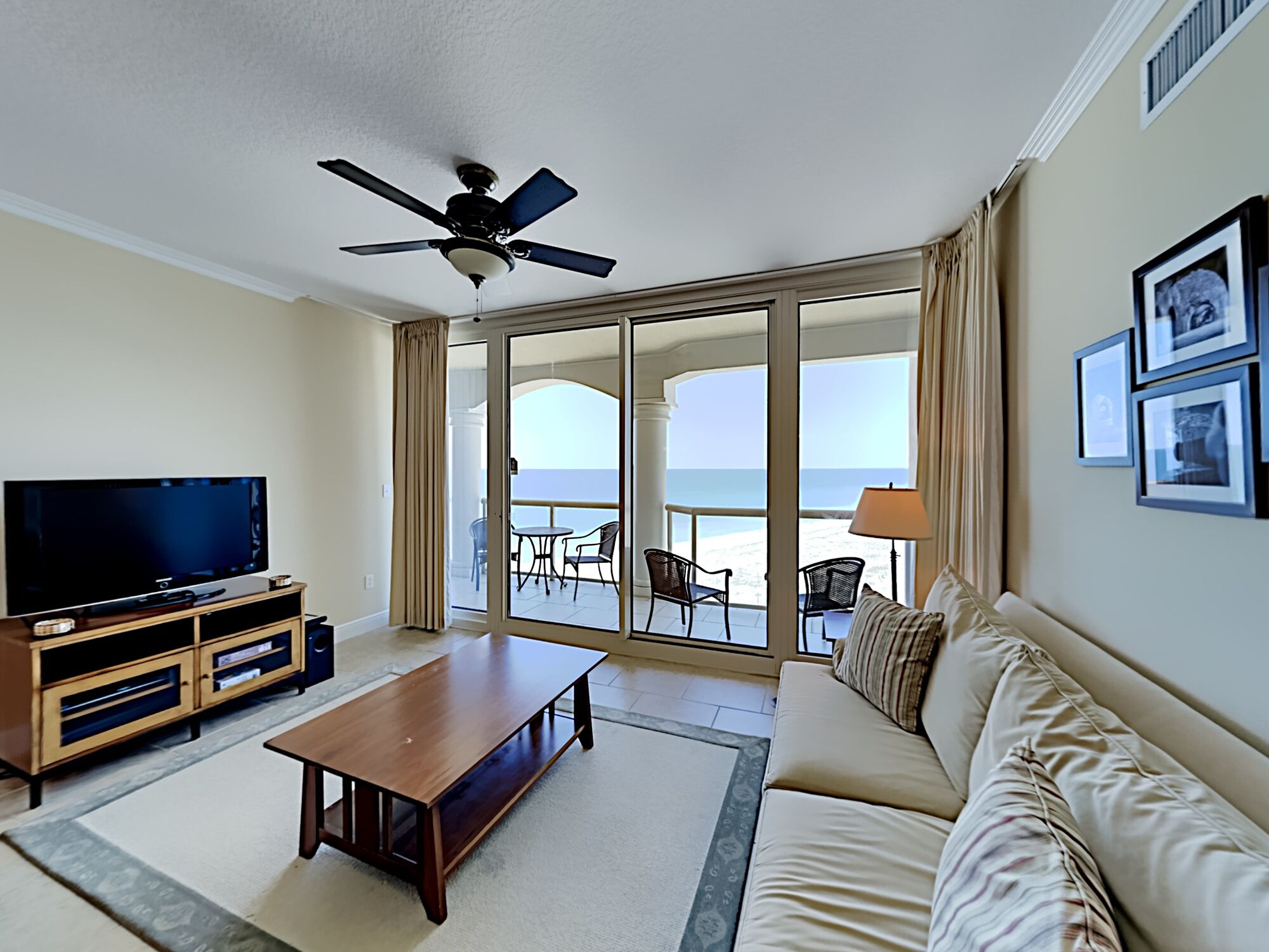 A living room with a television, a wooden coffee table, a sofa, and a balcony with a view of the ocean.