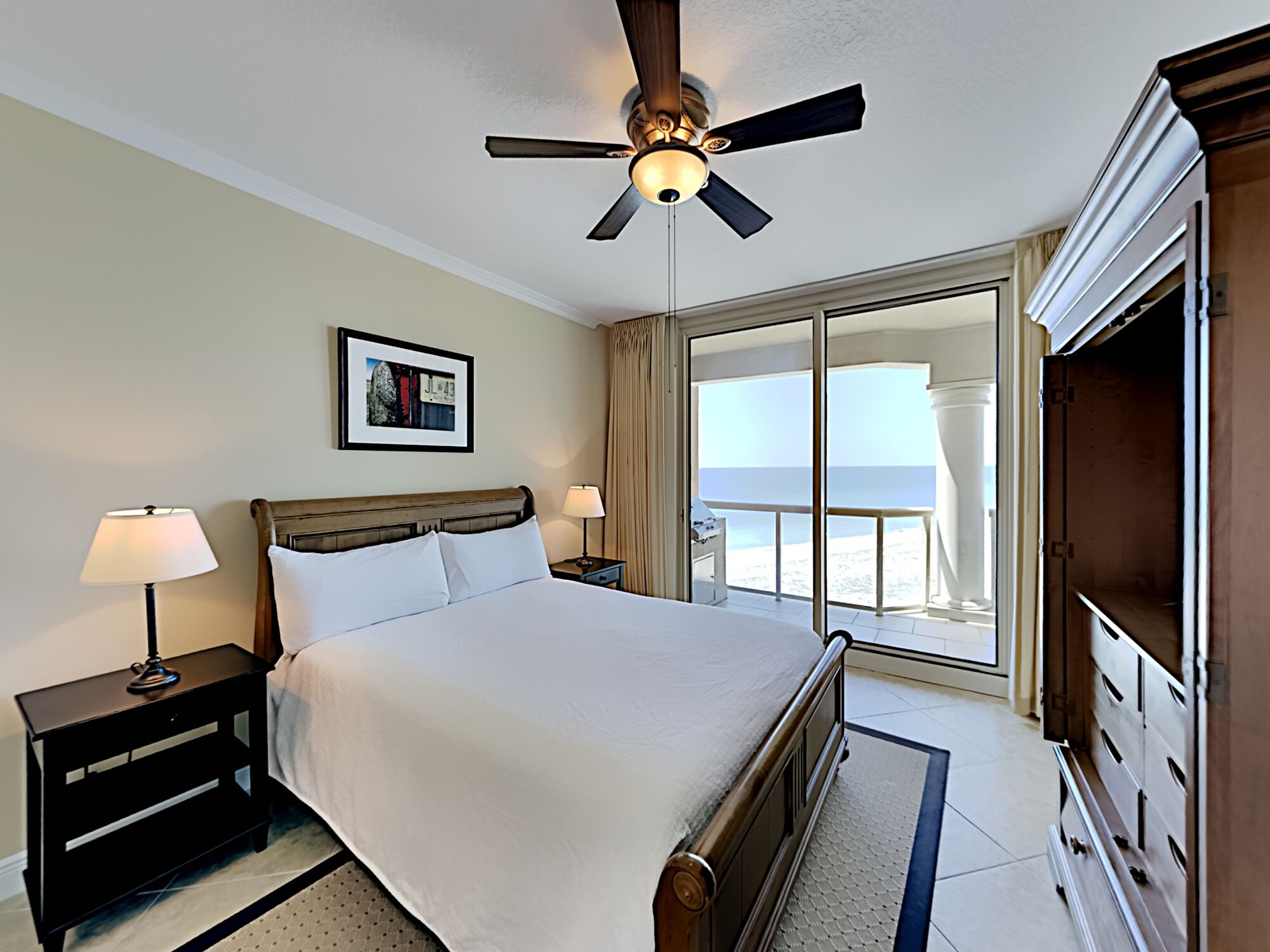 A hotel room with a bed, a ceiling fan, a nightstand with a lamp, a dresser, and a large window offering a view of the beach.