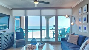 A living room with a large glass door leading to a balcony with a view of the beach. There is a blue sofa, a coffee table with a bowl of fruit, and a television on the left. - Portofino Island Resort 5 by Southern Vacation Rentals (Pensacola Beach)