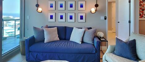 A living room with a blue sofa, a glass coffee table, and a grey armchair. There are framed pictures on the wall and a view of the outside.